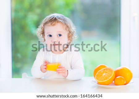 Happy little toddler girl drinking orange juice at a white table next to a big window