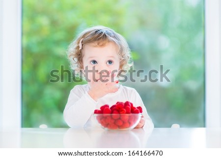 Cute Funny Toddler Girl Eating Raspberry At A White Table Next To A Big Window