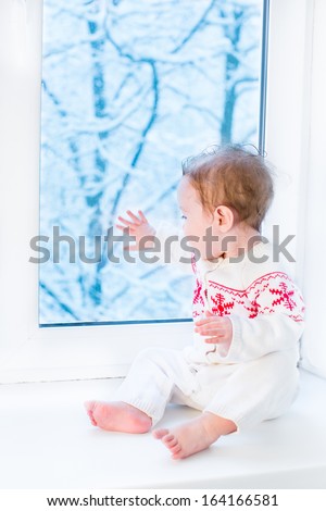 Cute Baby In A Knitted Sweater With Christmas Ornament Sitting At A Window Watching Snow Covered Trees