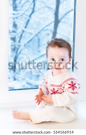 Funny little baby sitting next to a window to a garden with snow covered trees