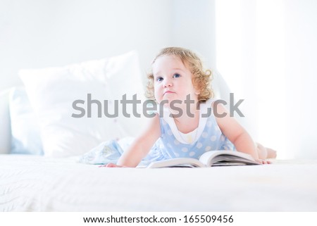 Funny toddler girl in a blue dress reading a book on a white bed