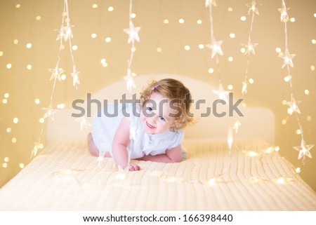 Beautiful curly toddler girl in a white dress jumping on a white bed between soft warm Christmas lights