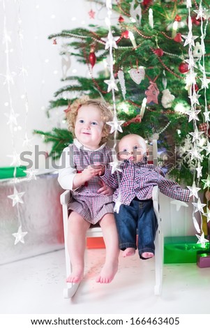 Cute laughing toddler girl and her little baby brother sitting together in a white rocking chair under a beautiful Christmas tree