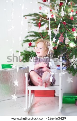 Happy laughing toddler girl playing with Christmas lights sitting in a white rocking chair under a decorated tree with presents