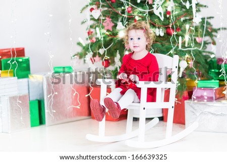 Cute curly toddler girl in a red dress relaxing in a white rocking chair under a Christmas tree between colorful present boxes