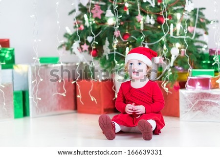 Cute little toddler girl in a Santa hat and red dress playing with a red toy bell sitting under a Christmas tree between colorful presents boxes