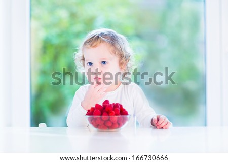 Funny toddler girl eating raspberries in the morning at a big window with garden view