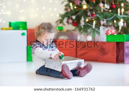 Funny little toddler girl opening her Christmas present under a beautiful Christmas tree