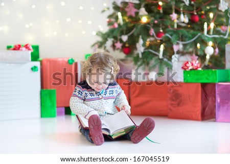 Little happy toddler girl with curly hair wearing a warm knitted jacket reading a book under a beautiful Christmas tree