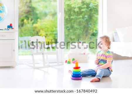 Smiling toddler girl playing with a colorful pyramid in a beautiful white room with a big window into the garden