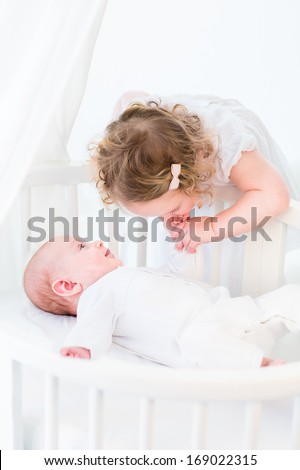 Cute toddler girl in a white dress kissing the hand of her baby brother relaxing in a white round crib