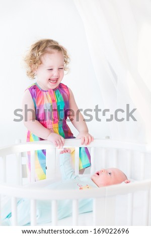 Adorable laughing toddler girl standing at a white round bed of her newborn baby brother
