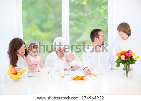 Happy young family with three children - teenager boy, cute toddler girl and a newborn baby - enjoying breakfast together with their grandmother in a sunny dining room with a big window