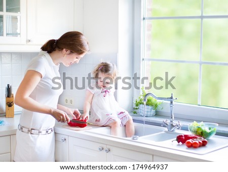 Cute toddler girl with curly hair wearing a white dress helping her mother to cook vegetables in a beautiful sunny kitchen with a big garden view window