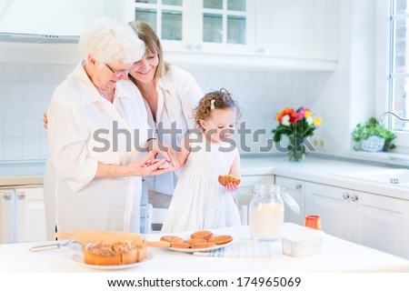Happy young woman baking a cake with her senior mother and cute toddler girl n a beautiful white kitchen