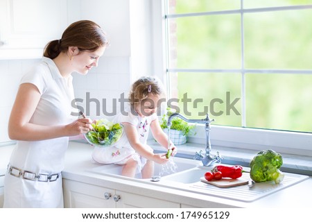 Young mother and her adorable toddler daughter cooking salad together in a beautiful white kitchen with a big garden view window