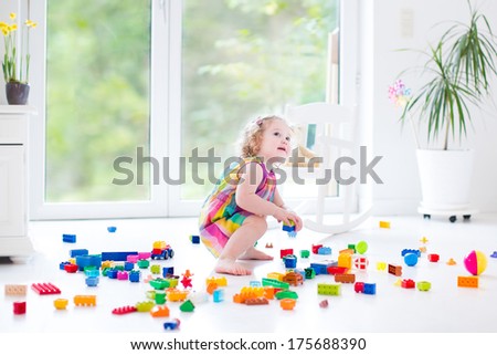 Sweet Curly Toddler Girl Playing With Colorful Blocks Sitting On A Floor In A Sunny Bedroom With A Big Window