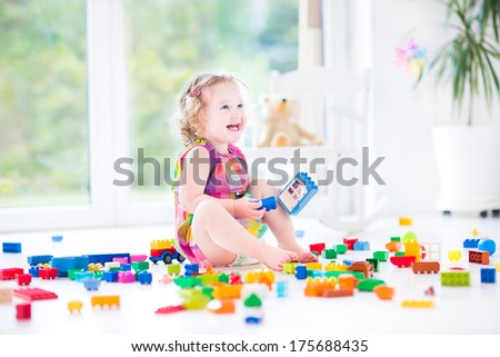 Adorable laughing toddler girl playing with colorful blocks sitting on a floor in a sunny bedroom with a big window