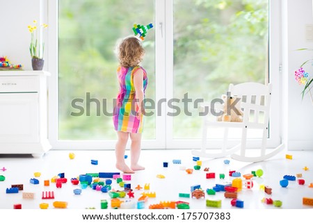 Cute laughing toddler girl playing with colorful blocks, building and airplane in a sunny bedroom with a big window