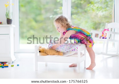 Cute curly toddler girl playing with her teddy bear in a sunny bedroom with big garden view windows