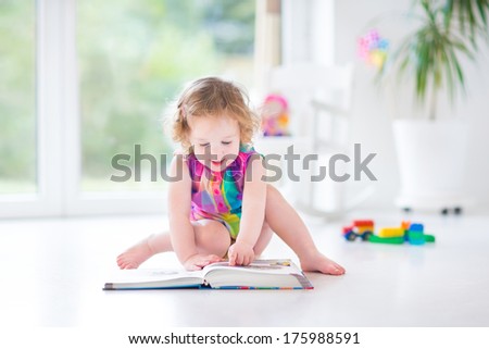 Funny Toddler Girl Reading A Book Sitting On A Floor In A White Room With Big Garden View Windows