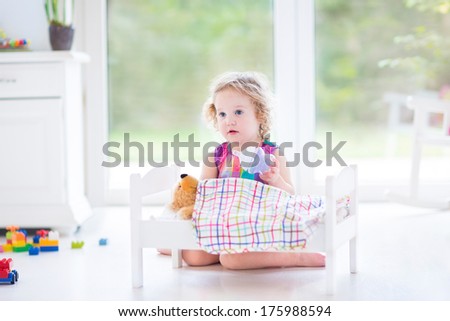 Funny toddler girl feeding her toy bear in a sunny room with big garden view windows