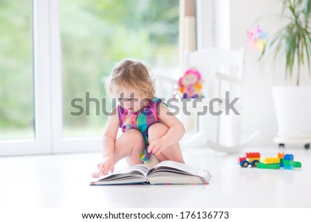 Adorable Toddler Girl Reading A Book In A Sunny Room With Big Windows