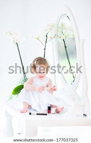 Adorable toddler girl with curly hair wearing a white dress playing with make up and cosmetics in front of a round mirror in a white sunny bedroom