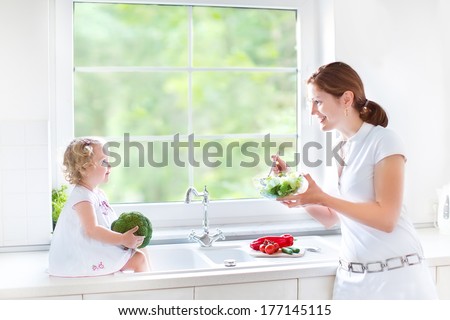 Young mother and her cute toddler daughter cooking together a healthy salad for lunch in a beautiful  white sunny kitchen with a big window