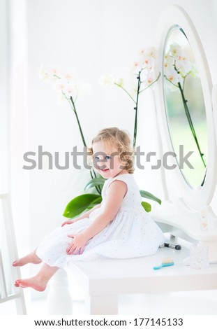 Pretty toddler girl with curly hair wearing a white dress playing with make up and cosmetics in front of a round mirror in a white sunny bedroom
