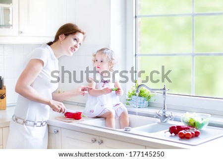 Young Mother And Her Adorable Curly Toddler Daughter Washing Vegetables Together In A Kitchen Sink Getting Ready To Make Salad For Lunch In A Sunny White Kitchen With A Big Garden View Window