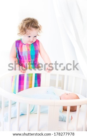Cute newborn baby boy looking at his toddler sister standing at his white round crib on a sunny morning