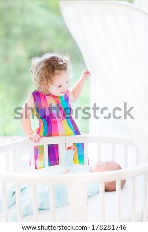 Cute newborn baby boy looking at his toddler sister standing at his white round crib on a sunny morning next to a big garden view window
