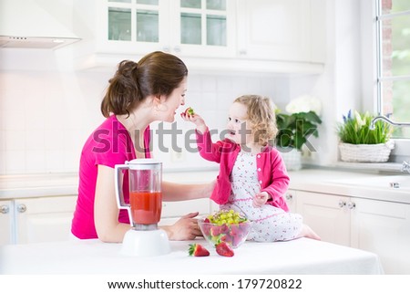 Happy laughing toddler girl and her beautiful young mother making fresh strawberry and other fruit juice for breakfast together in a sunny white kitchen with a window