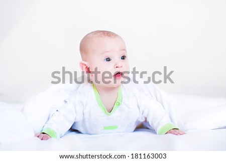 Adorable funny laughing baby playing peek-a-boo under a white blanket in a sunny bedroom waking up in the morning