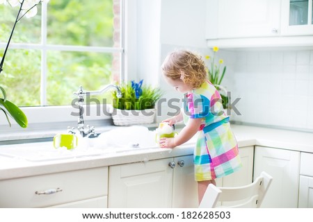 Cute curly toddler girl in a colorful dress washing dishes, cleaning with a sponge and playing with foam in the sink in a beautiful sunny white kitchen with a garden view window in a modern home