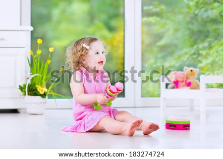 Cute curly laughing toddler girl in a pink dress playing tambourine and maracas in a sunny room with a big garden view window with a toy bed and spring flowers next to her, white modern interior home
