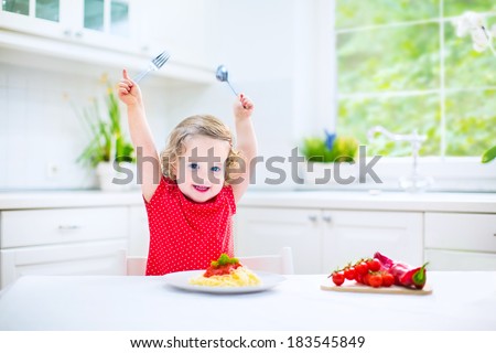Cute curly laughing toddler girl in a red shirt playing with fork and spoon eating spaghetti with tomato sauce and vegetables for healthy lunch sitting in a white sunny modern kitchen with big window