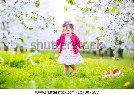 Adorable happy toddler girl with curly hair and flower crown wearing a red dress enjoying picnic in a beautiful blooming fruit garden with white blossoms on apple trees eating healthy snack