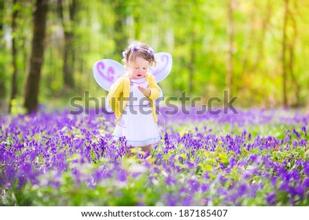 Adorable toddler girl with curly hair wearing a fairy costume with purple wings and yellow dress is playing in a beautiful spring forest with fresh blooming bluebell flowers on a sunny day in Germany