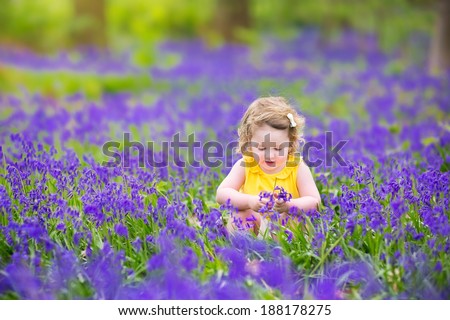 Adorable toddler girl with curly hair wearing a yellow dress playing with purple bluebell flowers in a sunny spring forest on a warm evening with beautiful sunset