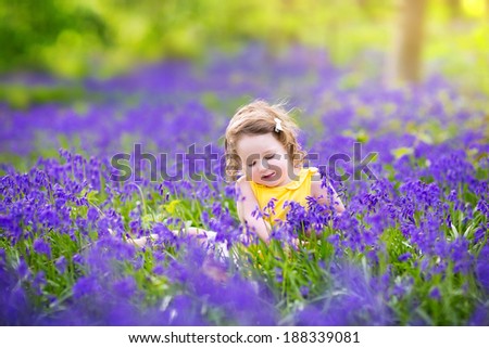 Adorable toddler girl with curly hair wearing a yellow dress playing with purple bluebell flowers in a sunny spring forest on a warm evening with beautiful sunset
