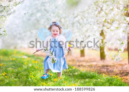 Adorable toddler girl with curly hair and flower crown wearing a magic fairy costume with a blue dress and angel wings playing in a sunny blooming fruit garden with cherry blossom and apple trees