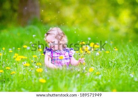 Adorable toddler girl with curly hair wearing a pink dress playing with yellow dandelions in a blooming spring garden on a sunny summer day
