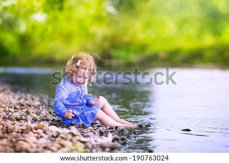 Adorable curly toddler girl wearing a blue dress playing at a river shore throwing stones into the water on a hot sunny summer day