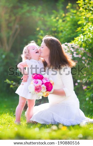 Beautiful young mother nd her adorable little daughter, cute little curly girl in a white dress, playing in the garden, hugging and kissing, with a bunch of red and pink flowers on a sunny summer day
