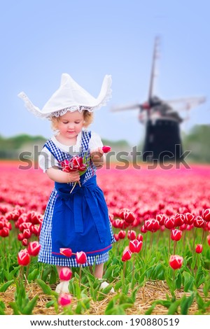 Adorable curly toddler girl wearing Dutch traditional national costume dress and hat playing in a field of blooming tulips next to a windmill in Amsterdam region, Holland, Netherlands