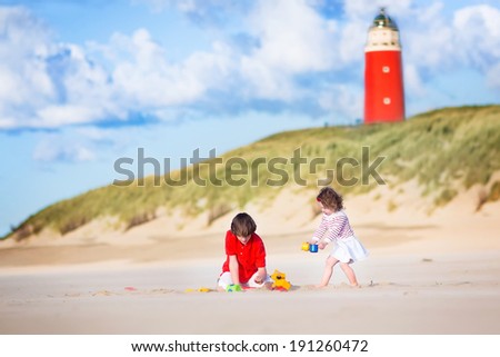 Happy children, young active boy and his adorable curly baby sister wearing a dress playing with sand toys on a sunny windy beach with a red lighthouse on Texel island, Holland, Netherlands
