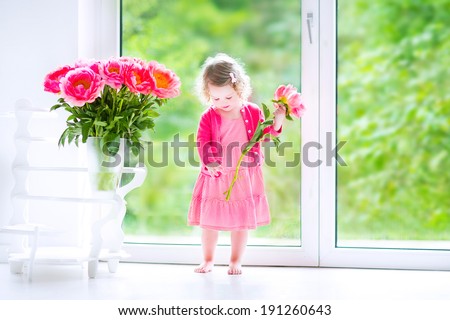 Cute happy toddler girl with curly hair wearing a pink dress playing with a bunch of beautiful big peony flowers in a vase in a white living room with big garden view windows