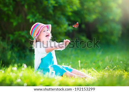 Happy laughing little girl wearing a blue dress and colorful straw hat playing with a flying butterfly having fun in the garden on a sunny summer day
